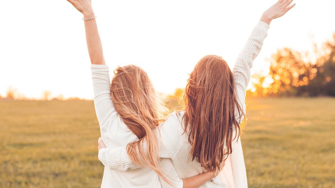 Zwei Frauen stehen Arm in Arm auf einem Feld im Sonnenuntergang und strecken ihre Arme in die Luft. | © Adobe Stock/Dima Aslanian