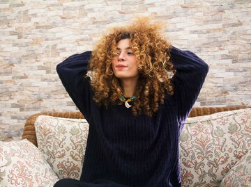 Junge Frau mit voluminösen Locken und dunklem Kaschmirpullover sitzt entspannt auf einem Sofa, hält die Hände im Haar und lächelt leicht. | © Getty Images/Hind Bouqartacha