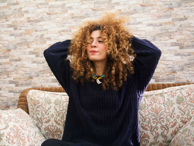 Junge Frau mit voluminösen Locken und dunklem Kaschmirpullover sitzt entspannt auf einem Sofa, hält die Hände im Haar und lächelt leicht. | © Getty Images/Hind Bouqartacha