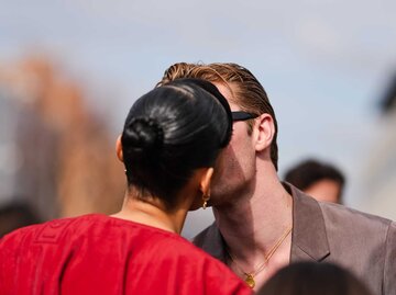 Eine Frau mit schwarzem Dutt und rotem Mantel gibt einem Mann in Sonnenbrille und braunem Blazer einen Kuss auf der Straße. | © Getty Images/Edward Berthelot