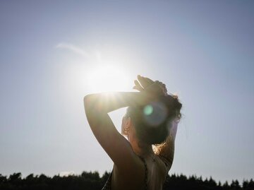 Eine junge Frau mit hochgesteckten Haaren steht in der Sonne, hebt die Hände über den Kopf und blickt in den strahlend blauen Himmel. | © Getty Images/Janina Steinmetz