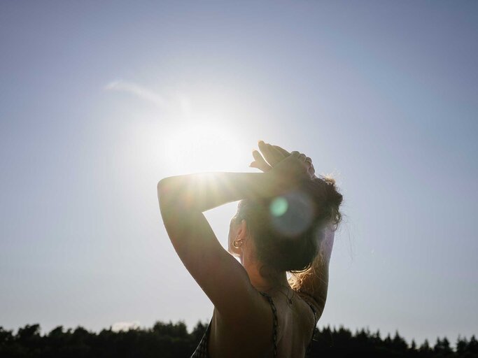 Eine junge Frau mit hochgesteckten Haaren steht in der Sonne, hebt die Hände über den Kopf und blickt in den strahlend blauen Himmel. | © Getty Images/Janina Steinmetz