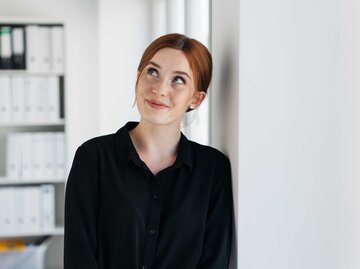 Rothaarige junge Frau trägt ein schwarzes Hemd und ist im Büro seitlich gegen die Wand gelehnt, schaut schräg nah oben. | © Adobe Stock/contrastwerkstatt