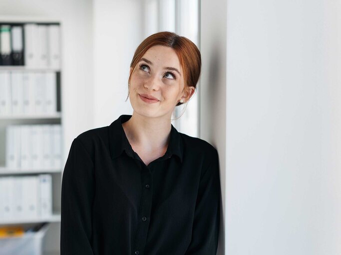 Rothaarige junge Frau trägt ein schwarzes Hemd und ist im Büro seitlich gegen die Wand gelehnt, schaut schräg nah oben. | © Adobe Stock/contrastwerkstatt