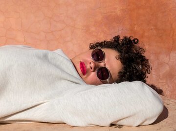 Frau mit Locken und Sonnenbrille liegt vor einer roten Wand auf dem Boden und schaut entspannt in die Kamera. | © Getty Images/Btihal Remli