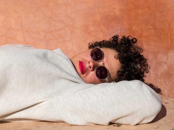 Frau mit Locken und Sonnenbrille liegt vor einer roten Wand auf dem Boden und schaut entspannt in die Kamera. | © Getty Images/Btihal Remli