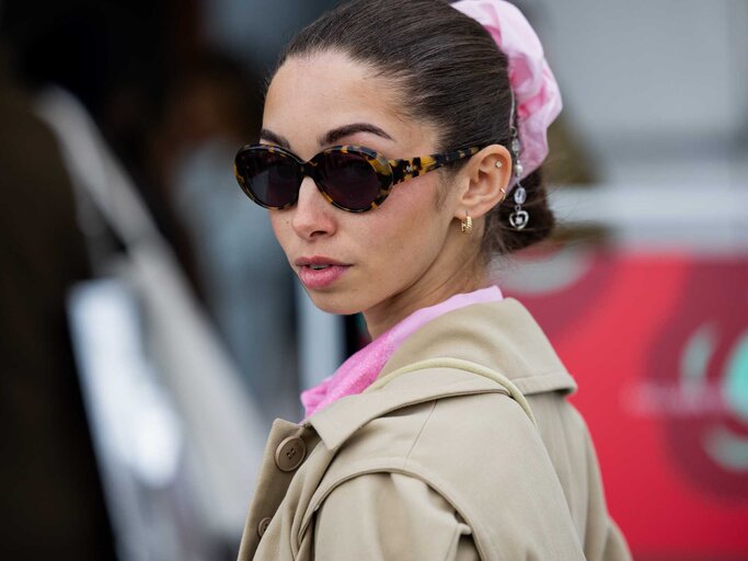 Junge Frau mit hochgestecktem Haar, rosa Haarband und goldenen Ohrringen blickt skeptisch mit dunkler Sonnenbrille und beigem Trenchcoat über die Schulter. | © Getty Images/Christian Vierig