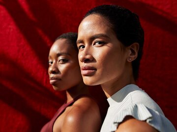 Zwei junge Frauen mit ernstem Blick stehen im Sonnenlicht vor einem roten Hintergrund, eine in Weiß, die andere in Bordeauxrot gekleidet. | © Getty Images/Klaus Vedfelt