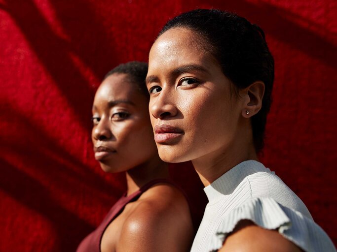 Zwei junge Frauen mit ernstem Blick stehen im Sonnenlicht vor einem roten Hintergrund, eine in Weiß, die andere in Bordeauxrot gekleidet. | © Getty Images/Klaus Vedfelt