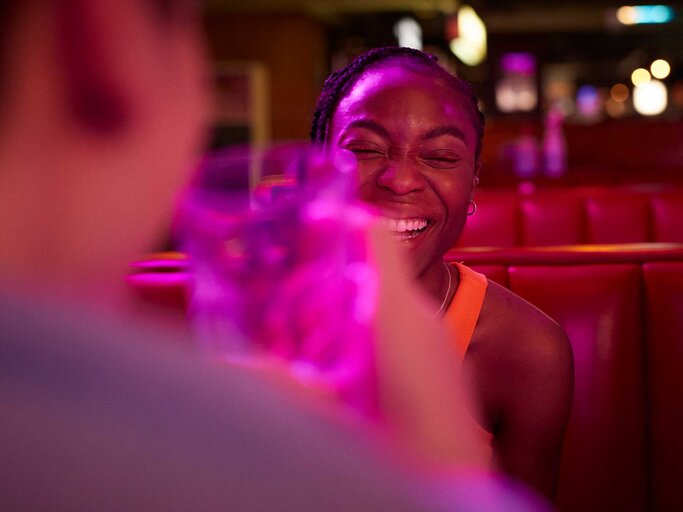 Frau sitzt bei einem Date in der Bar und lacht herzhaft, im Vordergrund verschwommen ihr Date mit einem Glas in der Hand.  | © Getty Images/Klaus Vedfelt