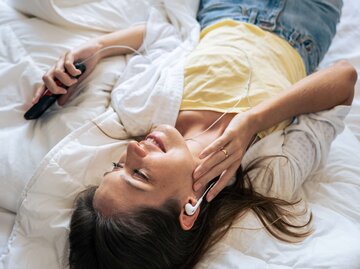 Eine Frau liegt auf dem Bett und hört Musik | © Adobe Stock/Kawee