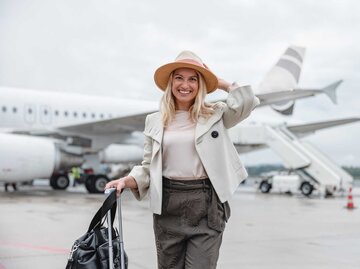 Lachende Frau auf dem Flugfeld mit Hut und Koffer. | © Getty Images/AzmanJaka