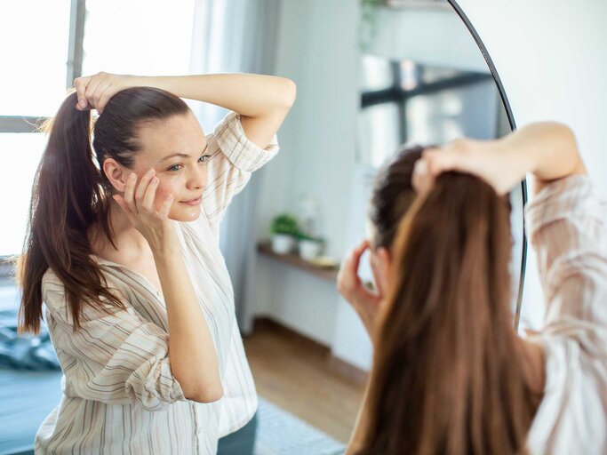 Frau mit braunen Haaren steht vor einem Spiegel und bindet sich einen hohen Pferdeschwanz. | © Adobe Stock/Vorda Berge