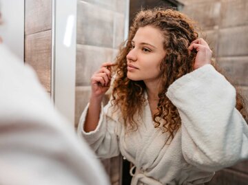 Frau mit Locken begutachtet ihre Haare im Spiegel.  | © Getty Images/urbazon