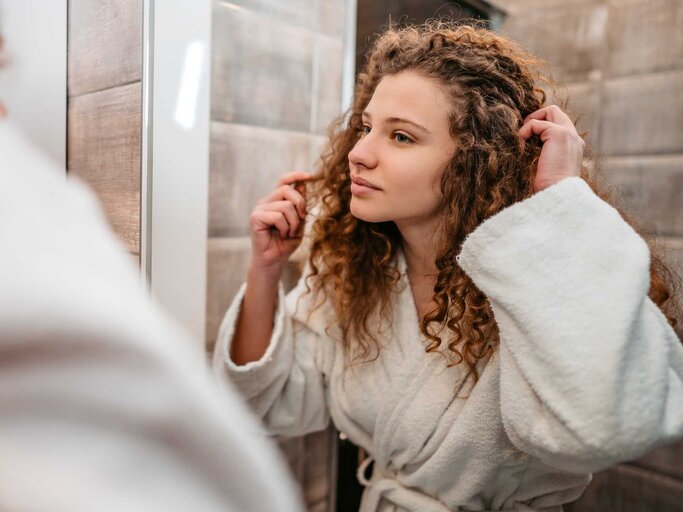 Frau mit Locken begutachtet ihre Haare im Spiegel.  | © Getty Images/urbazon