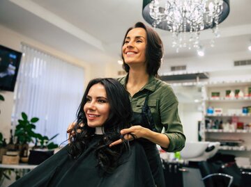 Eine lächelnde Frau mit dunklen, gelockten Haaren sitzt zufrieden im Friseursalon, während die Friseurin hinter ihr steht. | © Adobe Stock/My Ocean studio