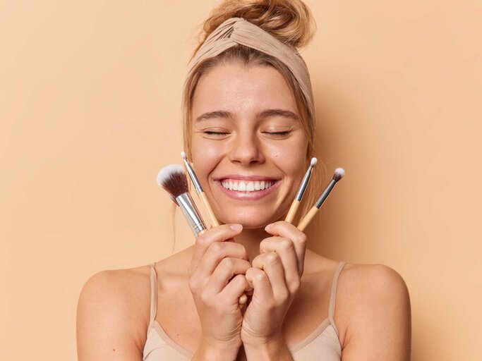 Frau hält sich Make-up Pinsel vor dem Gesicht und lächelt | © Adobe Stock/Prostock-studio