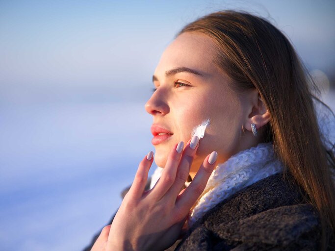 Junge Frau mit langen braunen Haaren trägt Creme auf ihre Wange auf, während sie im warmen Winterlicht in die Ferne blickt. | © Adobe Stock/Евгений Шемякин