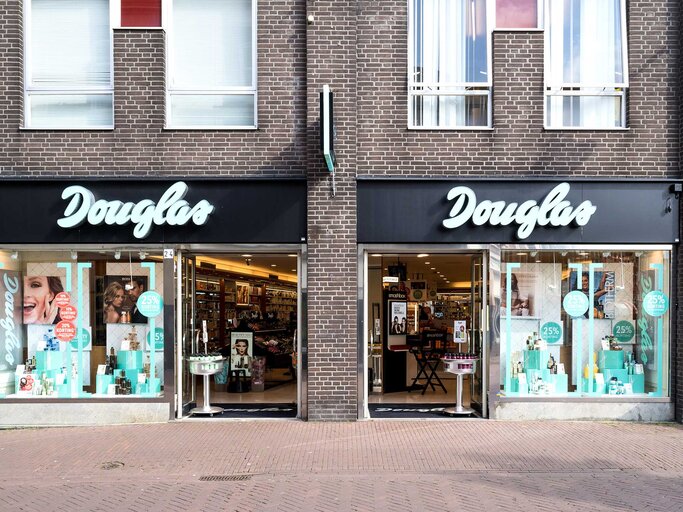 ALT-Text: "Die Vorderansicht eines Douglas-Kosmetikgeschäfts am Tag. Das Geschäft hat zwei große Schaufenster, die mit Werbung für Schönheitsprodukte und Angeboten dekoriert sind. In der Mitte befindet sich der Eingang, über dem in großen weißen Buchstaben 'Douglas' steht. | © Adobe Stock/Björn Wylezich