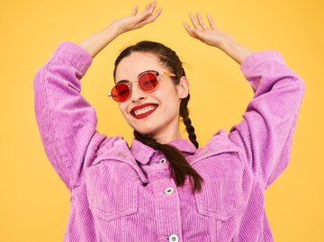 Eine junge Frau mit Lippenstift und roter Sonnebrille tanz lachend vor der Kamera | © Getty Images / Flashpop