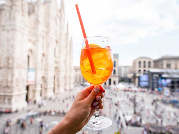Hand einer Frau hält ein Glas Aperol Spritz in die Luft - im Hintergrund ist die Kathedrale von Mailand. | © Adobe Stock/rh2010