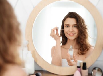 Frau steht mit Handtuch bekleidet im Badezimmer vor dem Spiegel und hält ein Serum für das Gesicht in der Hand, welches sie im Gesicht aufträgt. | © Adobe Stock/Prostock-studio