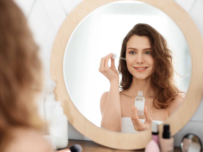 Frau steht mit Handtuch bekleidet im Badezimmer vor dem Spiegel und hält ein Serum für das Gesicht in der Hand, welches sie im Gesicht aufträgt. | © Adobe Stock/Prostock-studio