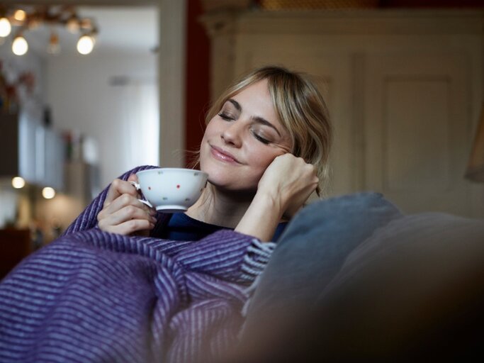 Junge Frau sitzt lächelnd auf einer Couch mit einer Tasse Tee | © GettyImages/Westend61