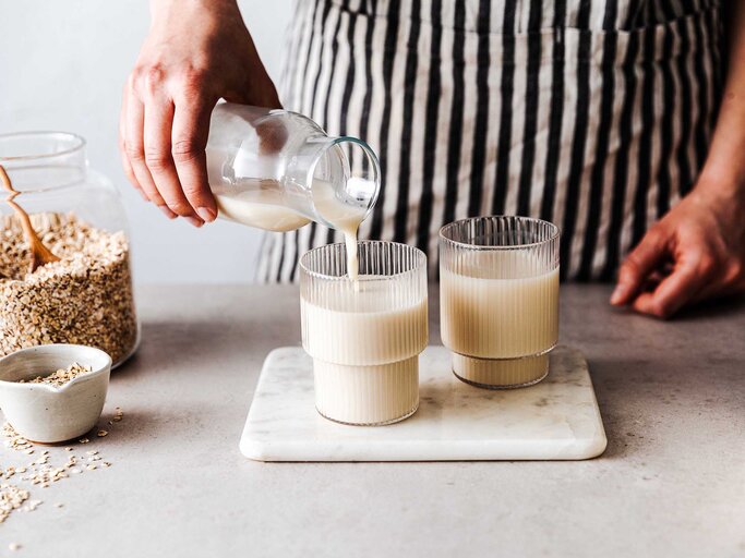 Frau mit Schürze schenkt am Küchentresen zwei Gläser Hafermilch ein. | © Getty Images / alvarez