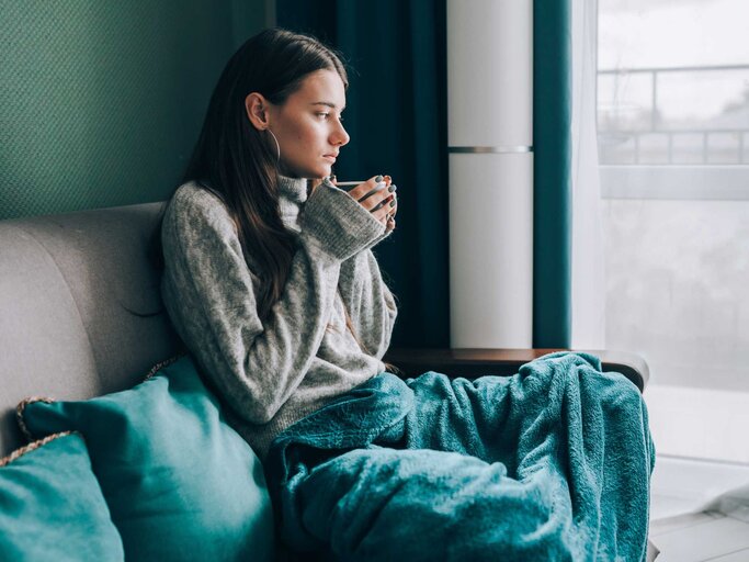 Frau sitzt mit einer Decke und Tasse in der Hand auf der Couch | © Getty Images/Olga Rolenko