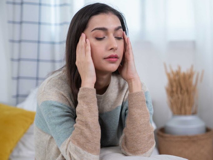 Frau sitzt auf ihrem Bett und fasst sich an die Schläfe | © Getty Images/Witthaya Prasongsin