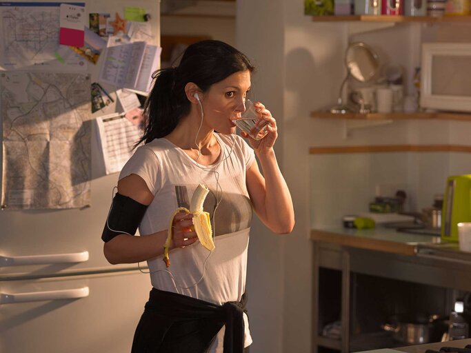 Frau steht morgens in der Küche und isst im Sportoutfit eine Banane und trinkt ein Glas Wasser. | © Getty Images / 10'000 Hours