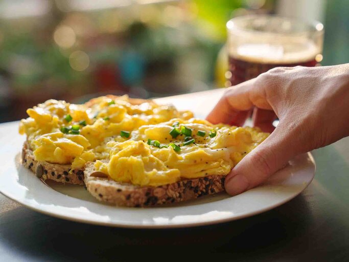 Hand greift nach einer Scheibe Brot, die üppig mit Rührei belegt ist | © Getty Images/Thai Liang Lim