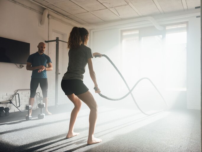 Eine Frau trainiert barfuß in einem Fitnessstudio mit Battle Ropes. | © Getty Images / Galdric