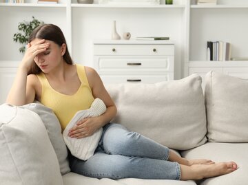 Frau sitzt mit Periodenschmerzen und einer Wärmflasche auf der Couch | © Adobe Stock/New Africa