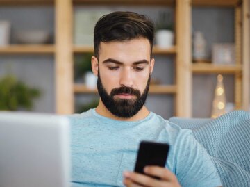 Junger Mann, der einen Laptop und sein Telefon benutzt, während er auf dem Sofa im Wohnzimmer sitzt. | © Getty Images/Aja Koska