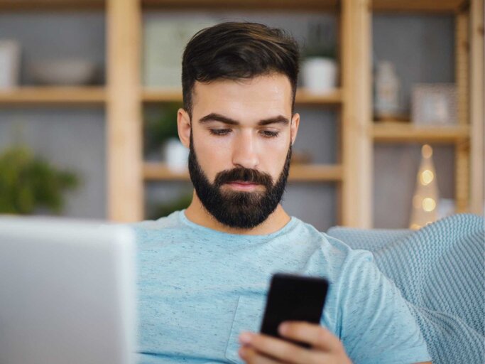 Junger Mann, der einen Laptop und sein Telefon benutzt, während er auf dem Sofa im Wohnzimmer sitzt. | © Getty Images/Aja Koska