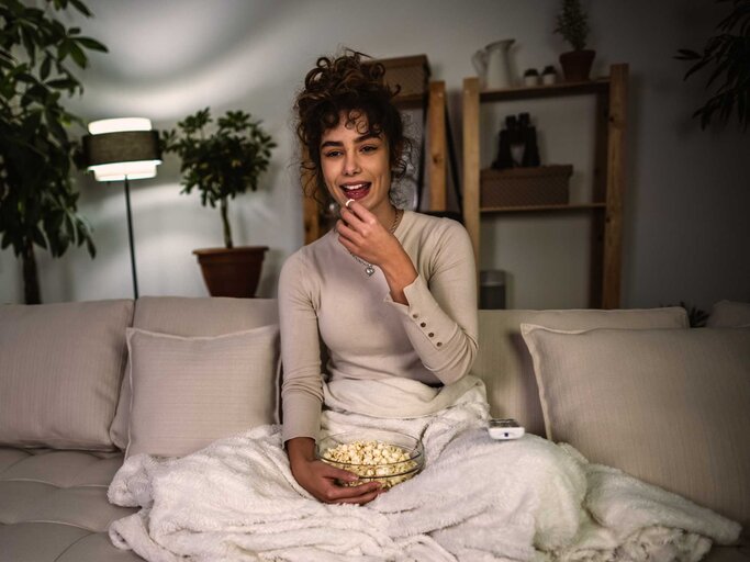 Junge Frau sitzt mit Decke und Popcorn auf der Couch. | © Getty Images/MilosStankovic