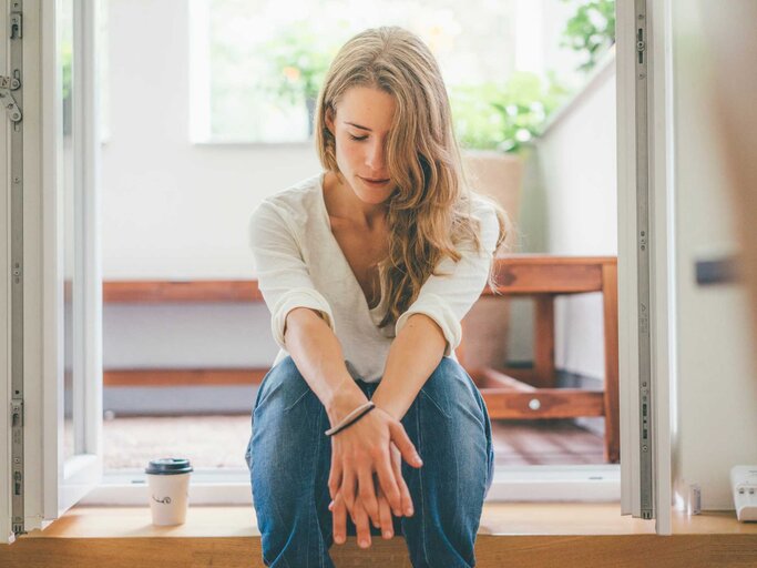 Junge Frau sitzt mit gesenktem Blick auf einer Stufe ihrer Wohnung. | © Getty Images/Westend61