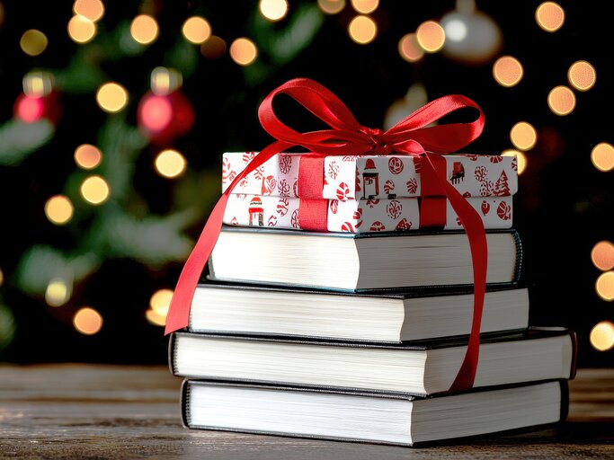 Ein Buchstapel mit einem Geschenk obendrauf vor einem Weihnachtsbaum. | © Adobe Stock/chayantorn/Generiert mit KI