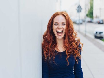 Frau mit roten langen Haaren steht an eine Wand gelehnt und lacht glücklich. | © Adobe Stock/contrastwerkstatt