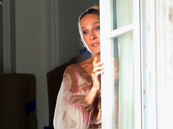 Sarah Jessica Parker steht bei den Dreharbeiten zu "And Just Like That" am Fenster und versteckt sich hinter dem Vorhang. | © Getty Images/Jose Perez/Bauer-Griffin / Kontributor