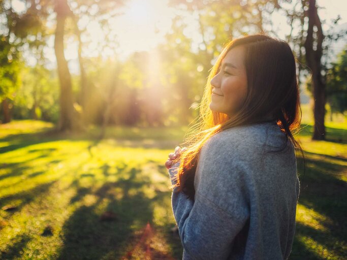 Glückliche Frau steht im Wald im Gegenlicht der Sonne. | © Adobe Stock/Farknot Architect