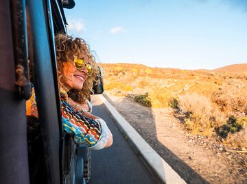 Junge Frau sitzt lachend im Auto und lehnt sich aus dem Fenster. | © Adobe Stock/simona