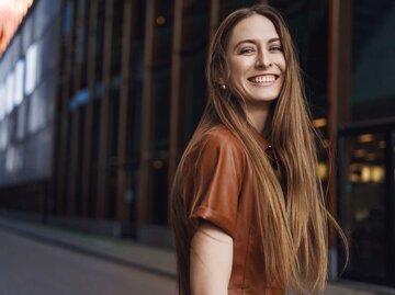 Junge Frau mit langem, braunem Haar und braunem Lederhemd lacht fröhlich und selbstbewusst auf einer leeren Stadtstraße in die Kamera. | © Adobe Stock/Liubov Levytska