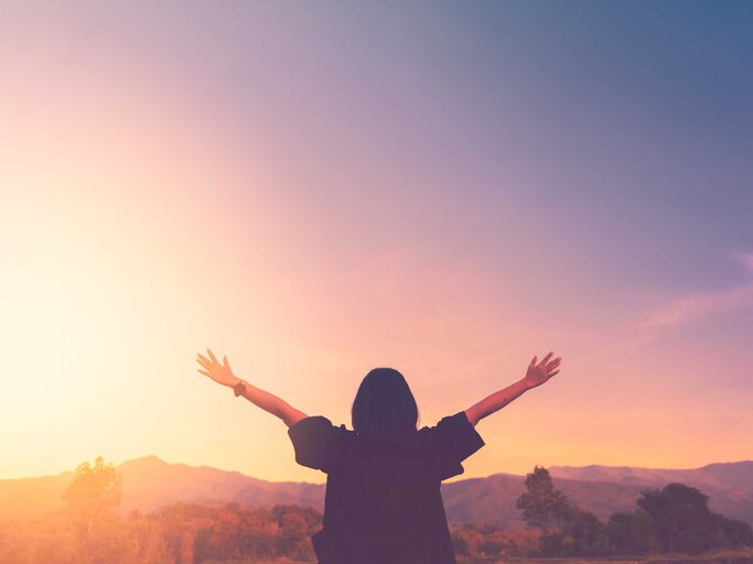 Eine Frau in dunkler Kleidung streckt bei Sonnenuntergang auf einem Hügel die Arme freudig in den Himmel. | © Adobe Stock/tonktiti