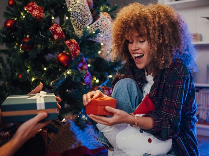 Frau sitzt vor dem Weihnachtsbaum und bekommt ein Geschenk | © Adobe Stock/Zamrznuti tonovi