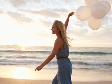 Lächelnde Frau hält Luftballons hoch und spaziert am Strand | © Adobe Stock/Jacob Lund