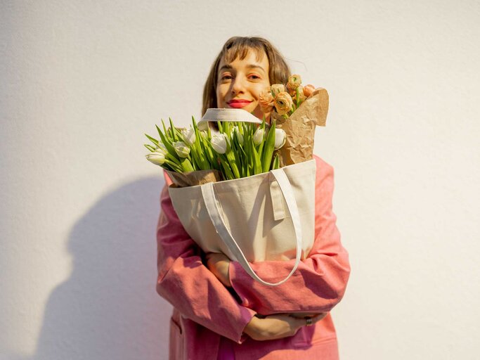 Lächelnde Frau in pinkem Outfit umarmt eine helle Stofftasche voller frischer Blumen, im Hintergrund eine weiße Wand mit Schatten. | © Adobe Stock/rh2010