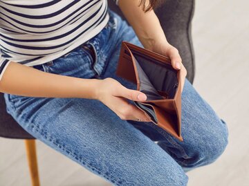 Frau mit Jeans und gestreiftem Oberteil sitzt auf einem Stuhl und hält einen offenen Geldbeutel in der Hand, in dem sich kein Geld befindet. | © Adobe Stock/Studio Romantic